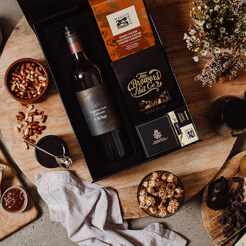 A gourmet gift box with a bottle of wine, nuts, crackers, and snacks, surrounded by small bowls of nuts, a wooden board, bread, chutney, a cloth napkin, and a bouquet of flowers on a rustic table.