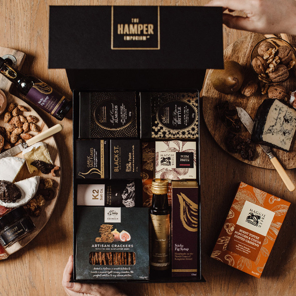 A person opens a luxury black hamper box filled with gourmet foods like crackers, fig syrup, and snacks. The box is on a wooden table with assorted cheeses, nuts, and a separate orange box of fruit paste nearby.