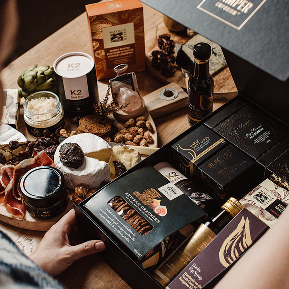 A person opens a gourmet gift box filled with assorted cheeses, crackers, snacks, and condiments, surrounded by a charcuterie board with meats, cheeses, and accompaniments on a wooden surface.