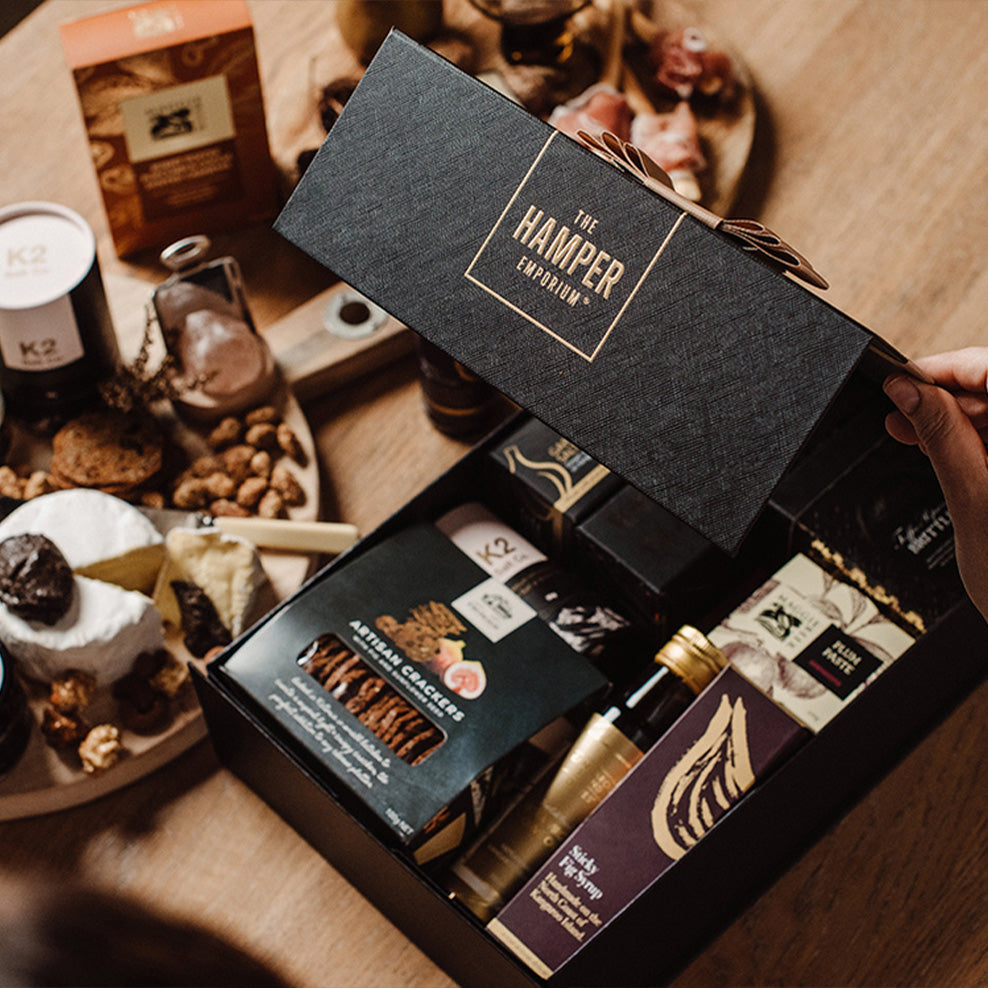 A person opens a black gift box labeled The Hamper Emporium, revealing assorted gourmet foods like crackers, nuts, and condiments. A wooden table with more snacks and cheeses is visible in the background.