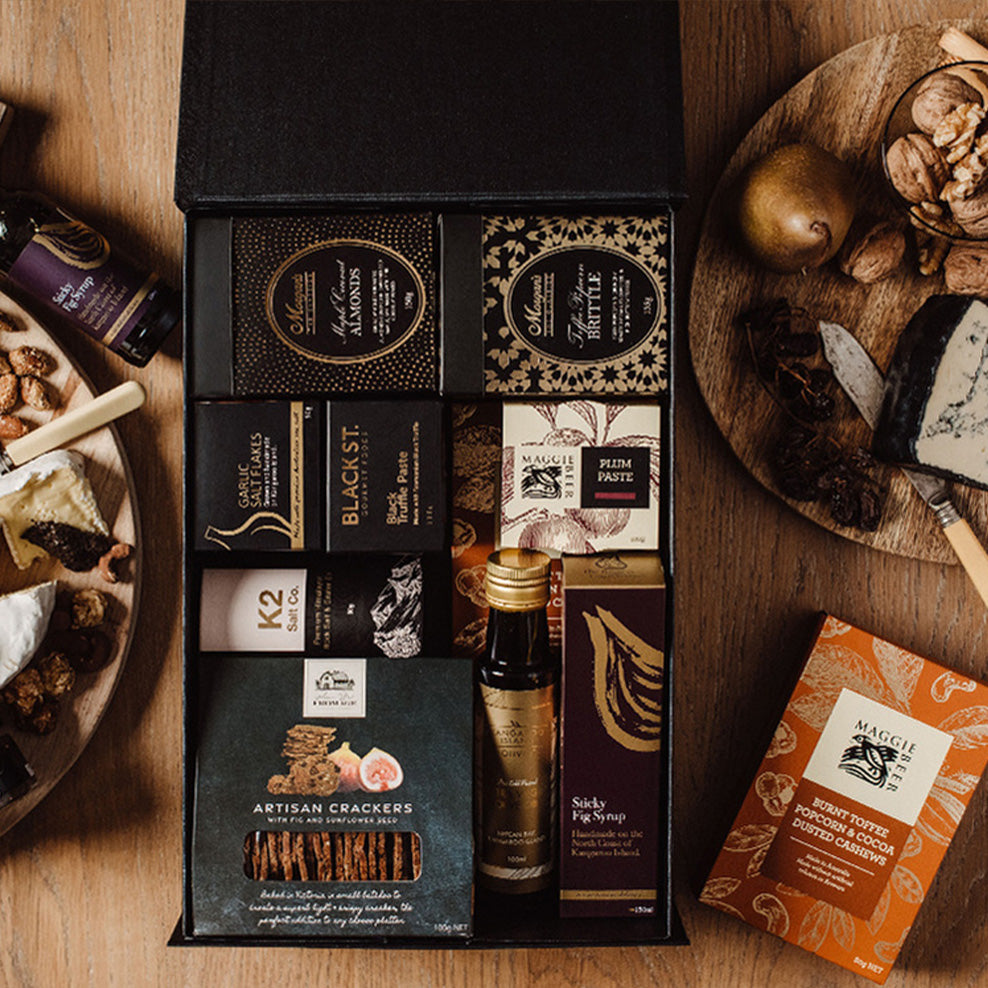 A gourmet gift box containing assorted chocolates, crackers, plum paste, oil, and other treats sits on a wooden table surrounded by cheese, nuts, and a cheese knife.