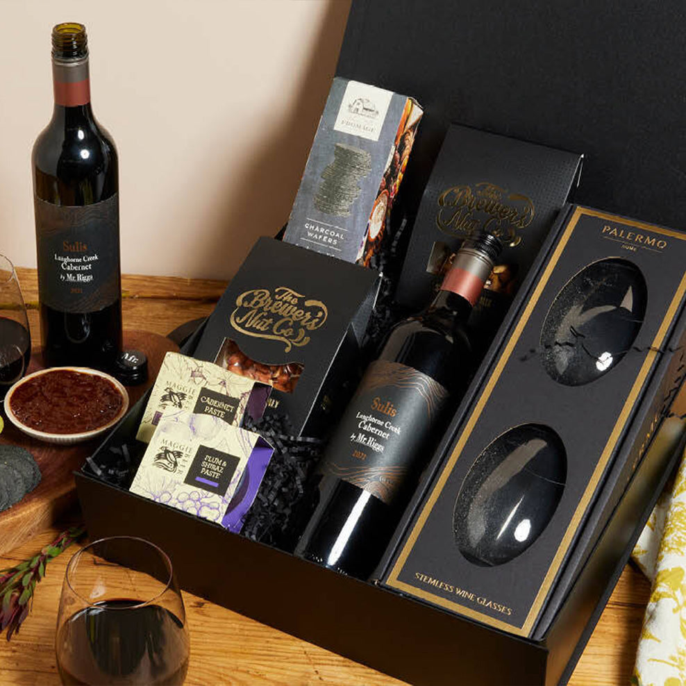 A gourmet gift box with a bottle of red wine, two stemless wine glasses, assorted crackers, chocolate, and cheese, arranged neatly on a wooden surface with a wine glass and bowl nearby.