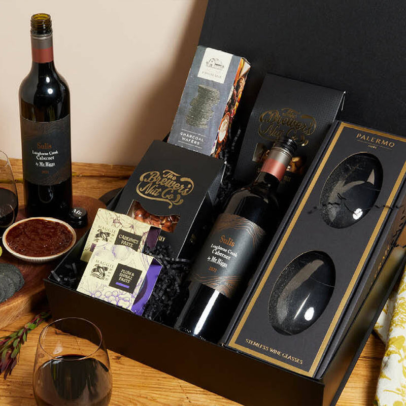 A gourmet gift box with a bottle of red wine, two stemless wine glasses, assorted crackers, chocolate, and cheese, arranged neatly on a wooden surface with a wine glass and bowl nearby.