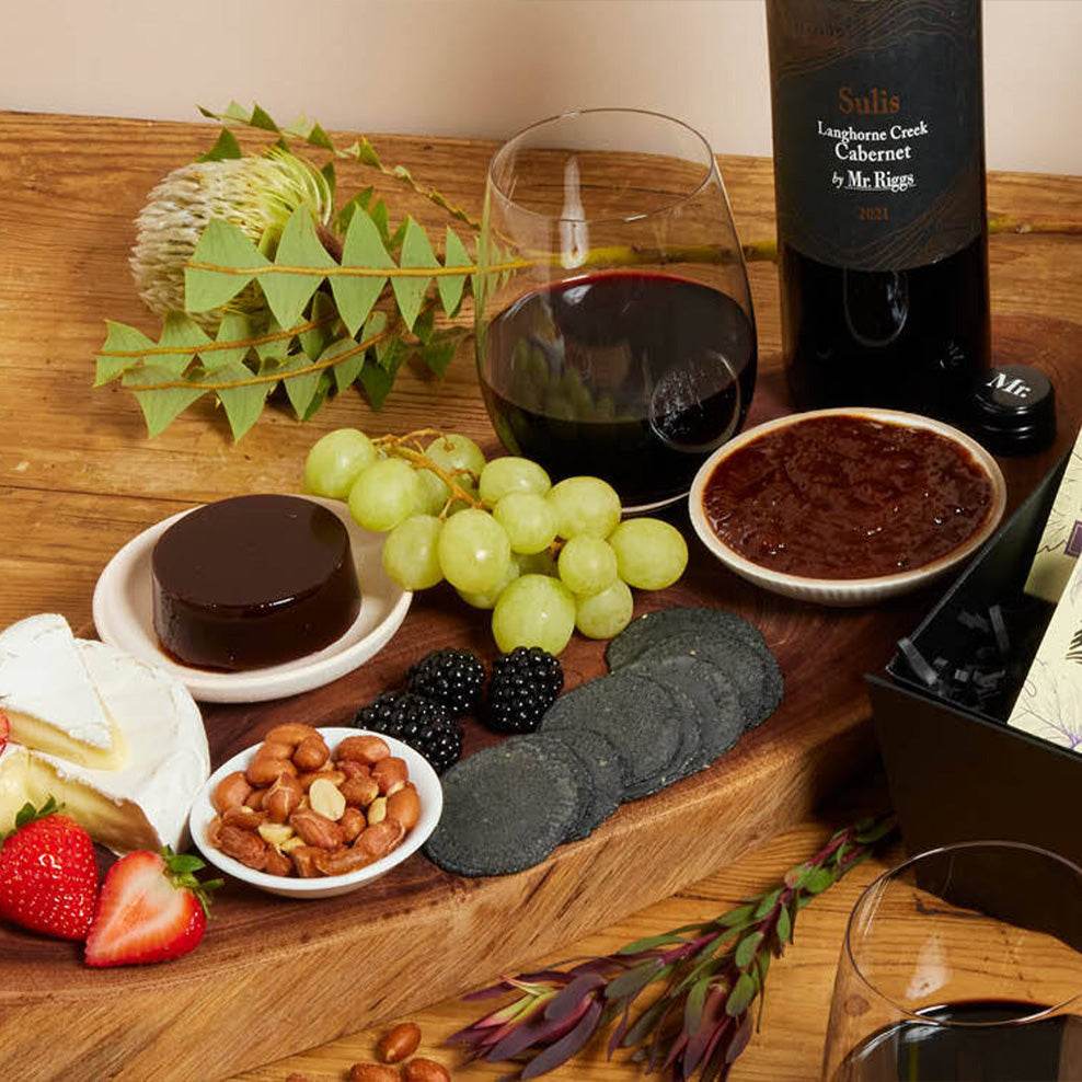 A wooden board with a bottle of red wine, two glasses of wine, grapes, blackberries, strawberries, mixed nuts, brie cheese, dark crackers, a chocolate dessert, and a dish of jam, decorated with leafy greenery.