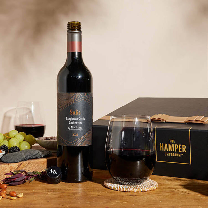 A bottle of red wine labeled Sulis Cabernet stands beside a filled wine glass, a black box labeled The Hamper Emporium, grapes, nuts, and a cork on a wooden surface.