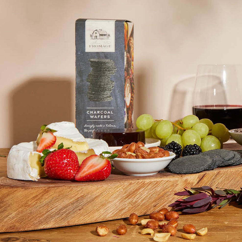 A wooden board with brie cheese, strawberries, grapes, blackberries, nuts, charcoal wafers, and a glass of red wine, with a box of charcoal wafers in the background.