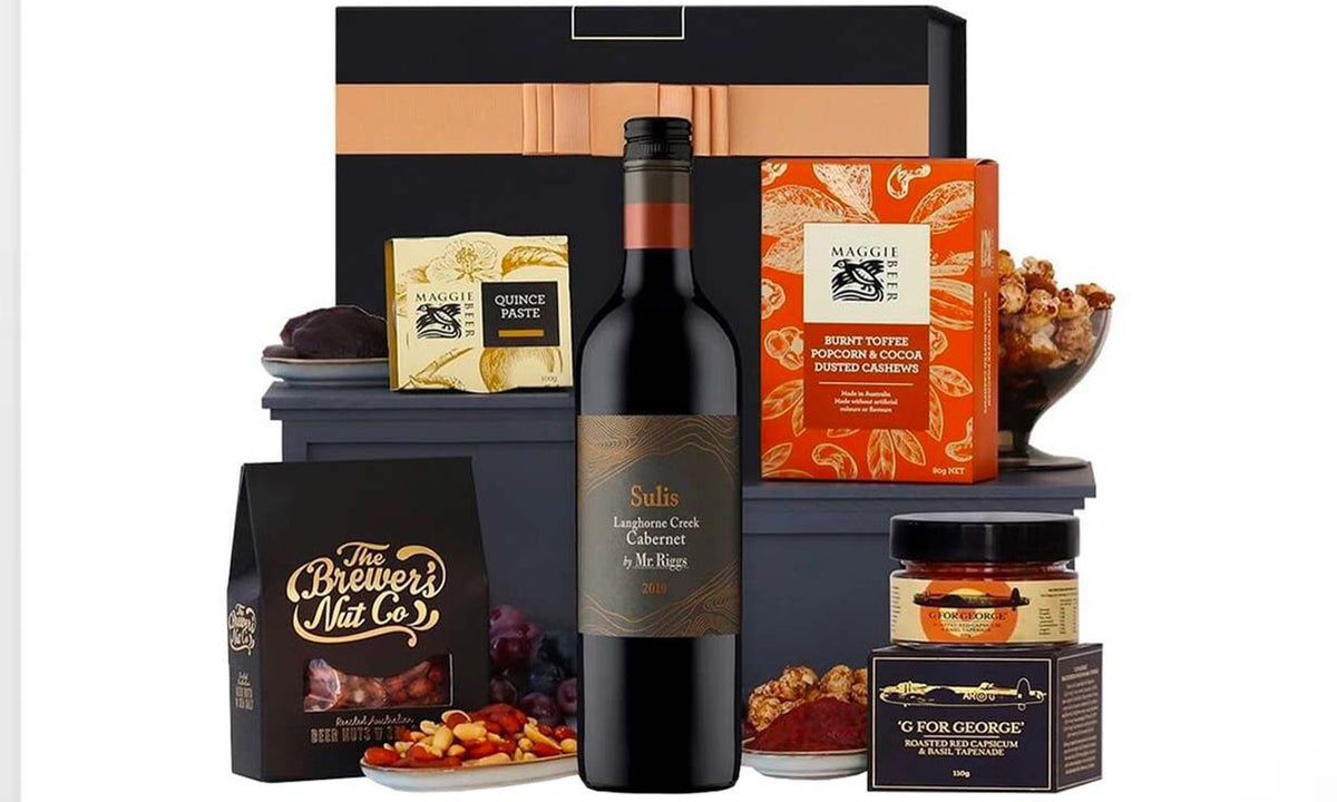 A gourmet gift hamper featuring a bottle of wine, assorted nuts, quince paste, crackers, a jar of relish, and a box of spiced cashews, all arranged around a black gift box.