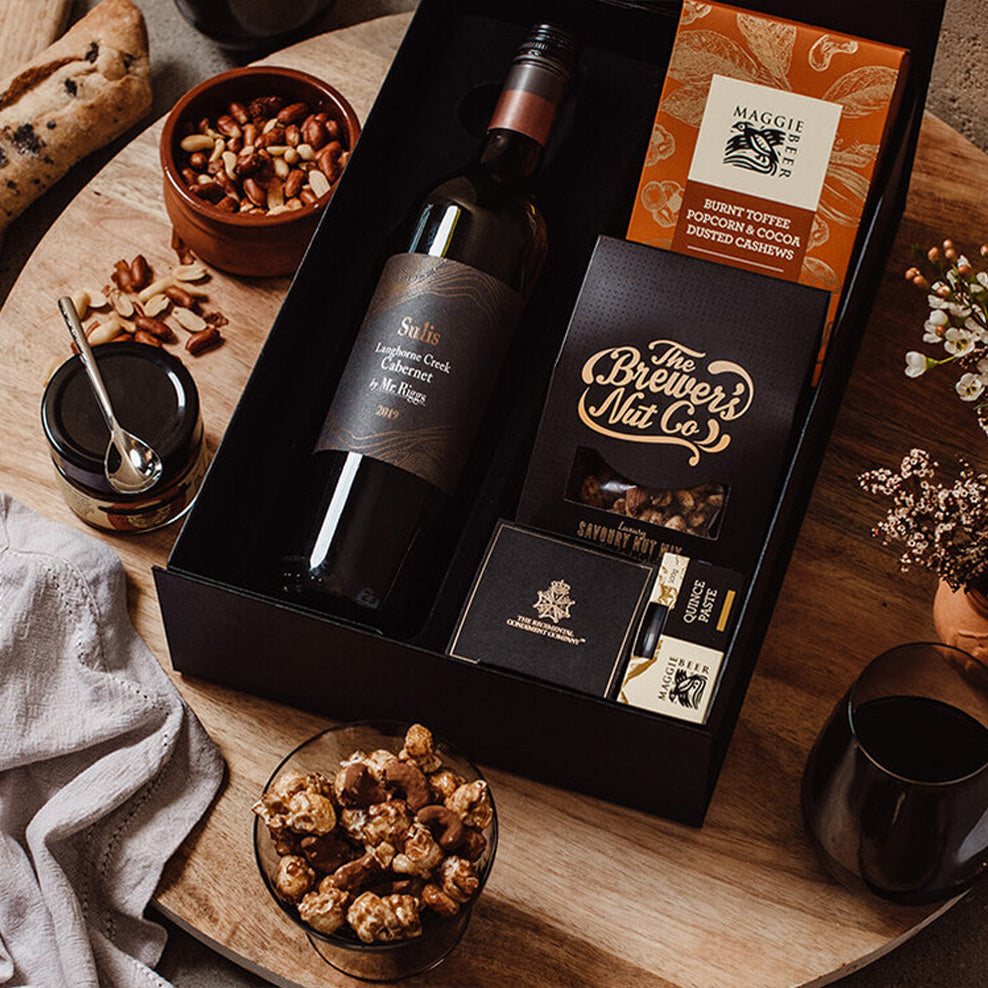 A gift box on a wooden table contains a bottle of red wine, nuts, chocolates, and gourmet snacks. Other snacks, a glass of wine, and a loaf of bread are arranged nearby for a cozy presentation.