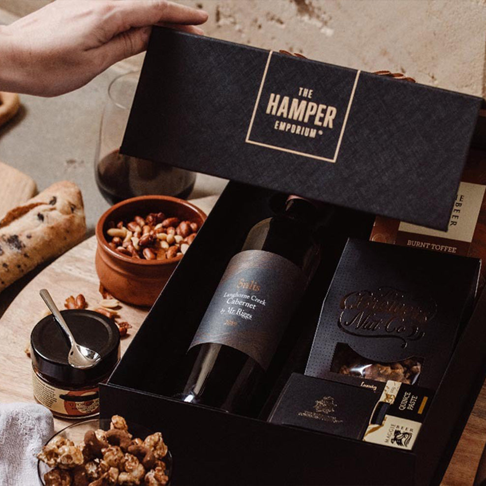 A hand opens a black gift box labeled The Hamper Emporium, revealing a bottle of wine, snacks, and treats. Surrounding the box are nuts, toffee, bread, a jar with a spoon, and a glass of red wine.
